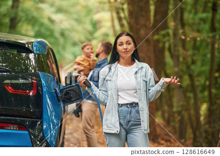 Woman holding wire. Mother, father and little son are waiting for electric car to charge outdoors Woman holding wire. Mother, father and little son are waiting for electric car to charge outdoors 128616649