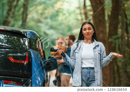 Woman holding wire. Mother, father and little son are waiting for electric car to charge outdoors Woman holding wire. Mother, father and little son are waiting for electric car to charge outdoors 128616650