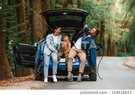 Conception of wellness. Mother, father and little son are waiting for electric car to charge outdoors Conception of wellness. Mother, father and little son are waiting for electric car to charge outdoors 128616651