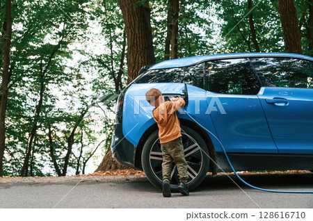 On the road. Cute young boy is charging the blue electric car outdoors On the road. Cute young boy is charging the blue electric car outdoors 128616710