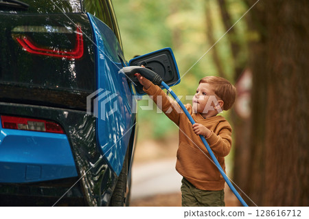 On the road. Cute young boy is charging the blue electric car outdoors 128616712