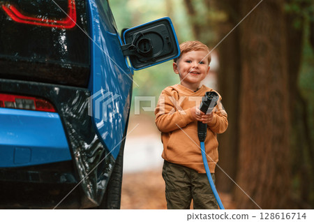 Standing and enjoying. Cute young boy is charging the blue electric car outdoors 128616714