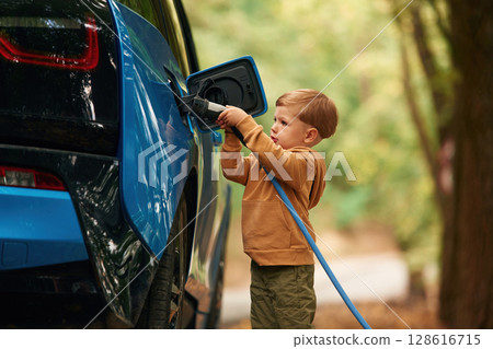 Standing and enjoying. Cute young boy is charging the blue electric car outdoors 128616715