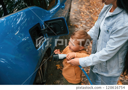 Learning the process. Mother with her little son are charging the electric car outdoors Learning the process. Mother with her little son are charging the electric car outdoors 128616754