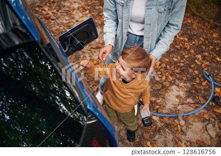 Learning the process. Mother with her little son are charging the electric car outdoors Learning the process. Mother with her little son are charging the electric car outdoors 128616756