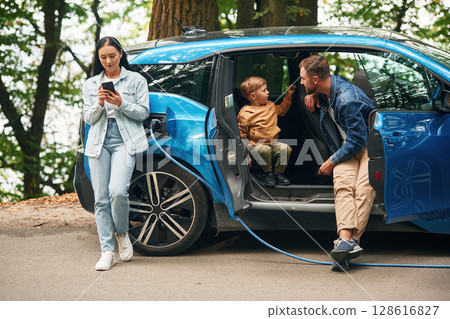 Road in woodland. Mother, father and little son are waiting for electric car to charge outdoors Road in woodland. Mother, father and little son are waiting for electric car to charge outdoors 128616827
