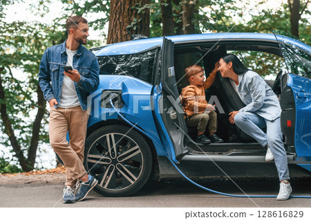 Road in woodland. Mother, father and little son are waiting for electric car to charge outdoors 128616829