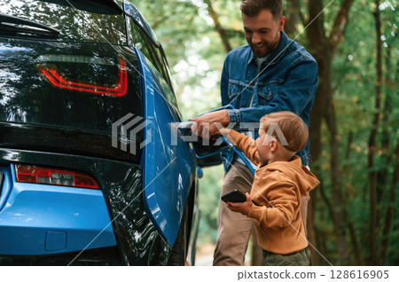 Teaching the boy where to put the wire. Father with his little son are waiting for electric car to charge Teaching the boy where to put the wire. Father with his little son are waiting for electric car to charge 128616905