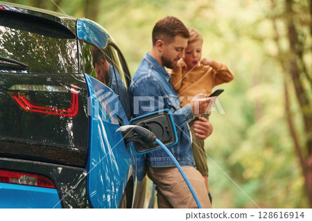 Holding boy in hands. Father with his little son are waiting for electric car to charge Holding boy in hands. Father with his little son are waiting for electric car to charge 128616914