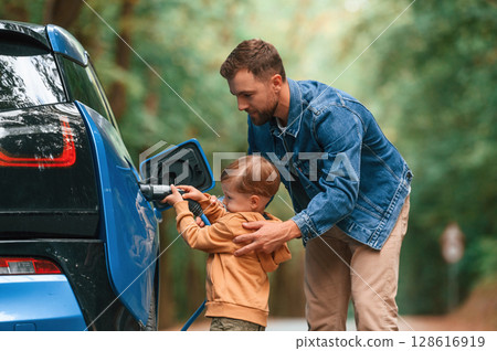 Teaching the boy where to put the wire. Father with his little son are waiting for electric car to charge Teaching the boy where to put the wire. Father with his little son are waiting for electric car to charge 128616919