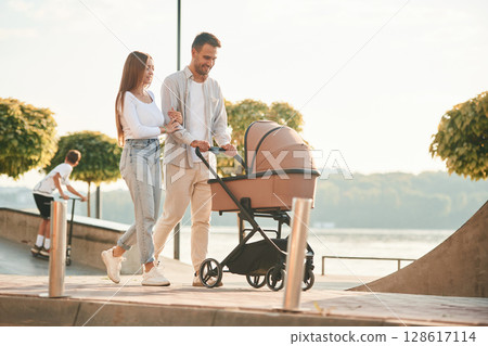 Little green trees behind. A young couple with a baby pram is walking together Little green trees behind. A young couple with a baby pram is walking together 128617114