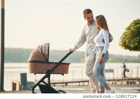 Embracing the woman by the waist. A young couple with a baby pram is walking together Embracing the woman by the waist. A young couple with a baby pram is walking together 128617124