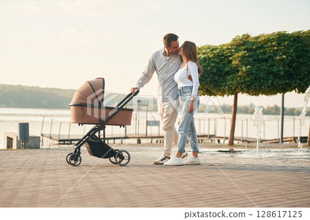 Embracing the woman by the waist. A young couple with a baby pram is walking together Embracing the woman by the waist. A young couple with a baby pram is walking together 128617125