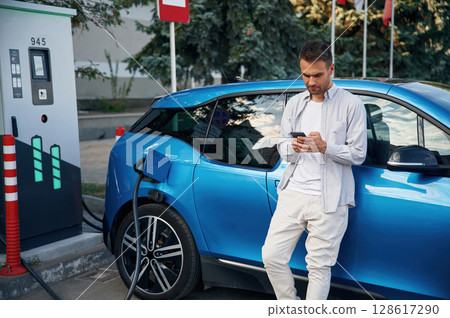 Standing with smartphone. Man with blue electric car on the charge station 128617290