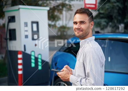 Handsome person with smartphone. Man with blue electric car on the charge station 128617292
