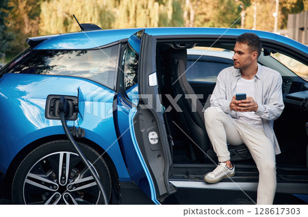 Handsome person with smartphone. Man with blue electric car on the charge station Handsome person with smartphone. Man with blue electric car on the charge station 128617303