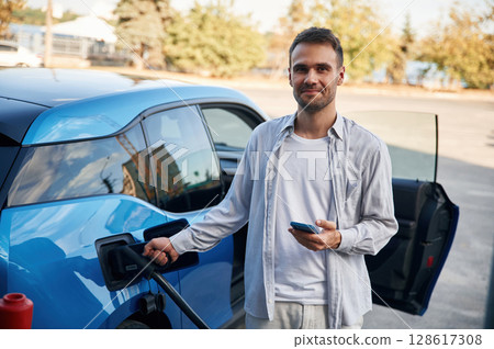 Front view. Man with blue electric car on the charge station 128617308