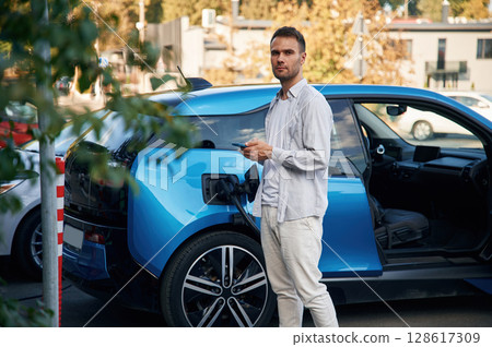 Front view. Man with blue electric car on the charge station Front view. Man with blue electric car on the charge station 128617309