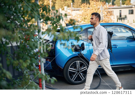 With backpack. Man with blue electric car on the charge station 128617311