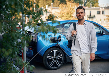 With backpack. Man with blue electric car on the charge station 128617313