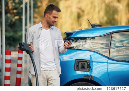 Wire in socket, holding smartphone. Man with blue electric car on the charge station Wire in socket, holding smartphone. Man with blue electric car on the charge station 128617317