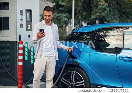 Wire in socket, holding smartphone. Man with blue electric car on the charge station 128617321