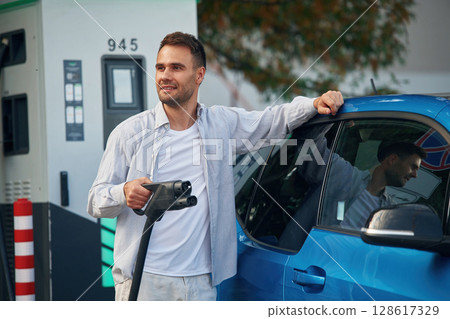 With wire in hand. Man with blue electric car on the charge station With wire in hand. Man with blue electric car on the charge station 128617329