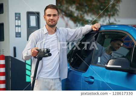 With wire in hand. Man with blue electric car on the charge station 128617330