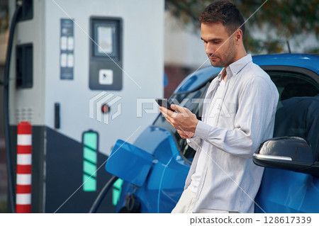 Side view, standing with smartphone, waiting. Man with blue electric car on the charge station Side view, standing with smartphone, waiting. Man with blue electric car on the charge station 128617339