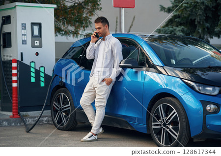 Conversation by phone. Man with blue electric car on the charge station 128617344