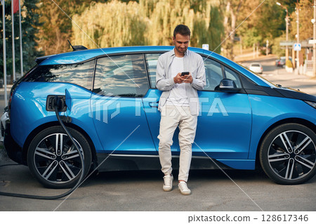 Blue vehicle, holding smartphone. Man with blue electric car on the charge station 128617346