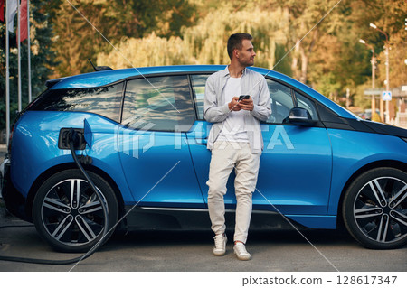 Blue vehicle, holding smartphone. Man with blue electric car on the charge station 128617347