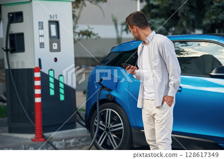 Blue vehicle, holding smartphone. Man with blue electric car on the charge station 128617349