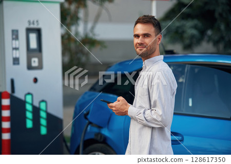 Blue vehicle, holding smartphone. Man with blue electric car on the charge station 128617350