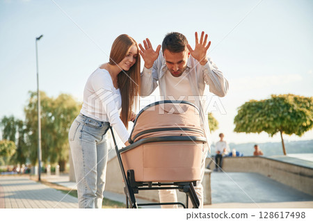 Man playing with kid, showing gestures. A young couple with a baby pram is walking together Man playing with kid, showing gestures. A young couple with a baby pram is walking together 128617498