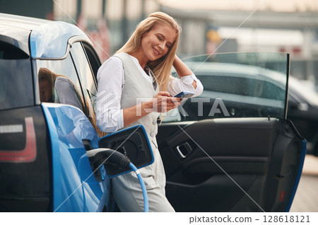 Smartphone in hands. Woman on the electric cars charge station at daytime Smartphone in hands. Woman on the electric cars charge station at daytime 128618121