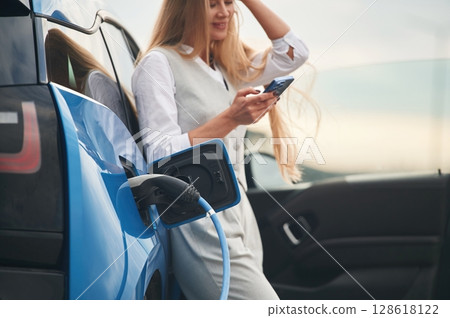 Smartphone in hands. Woman on the electric cars charge station at daytime Smartphone in hands. Woman on the electric cars charge station at daytime 128618122