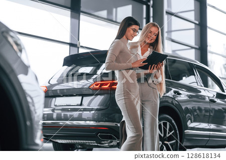 Standing and holding tablet. Female manager is helping woman customer in the car dealership salon Standing and holding tablet. Female manager is helping woman customer in the car dealership salon 128618134