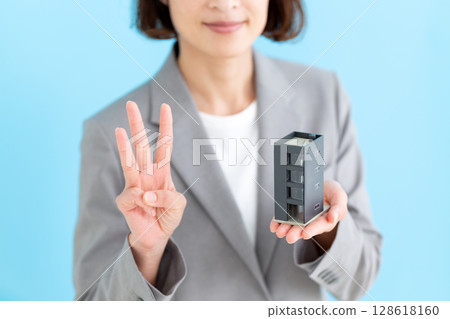 A middle-aged female business woman holding a model of an office building and raising three fingers A middle-aged female business woman holding a model of an office building and raising three fingers 128618160
