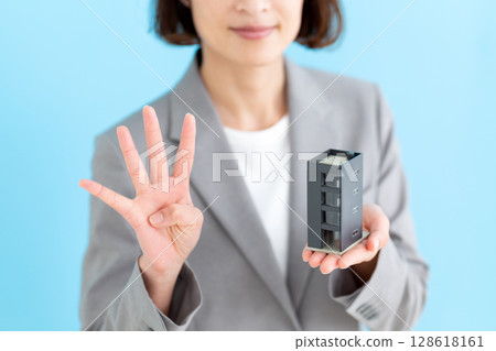 A middle-aged female business woman holding a model of an office building and showing four fingers 128618161