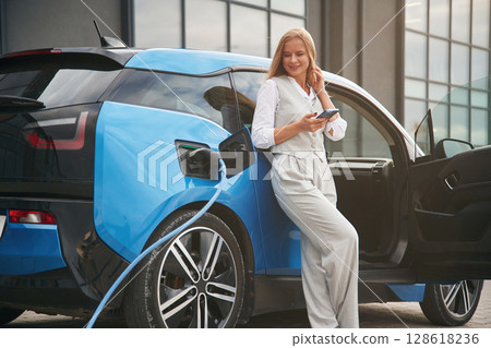 Holding smartphone. Woman on the electric cars charge station at daytime Holding smartphone. Woman on the electric cars charge station at daytime 128618236