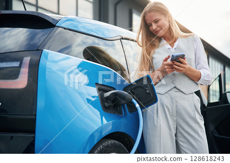 Wire is in the socket. Woman on the electric cars charge station at daytime 128618243