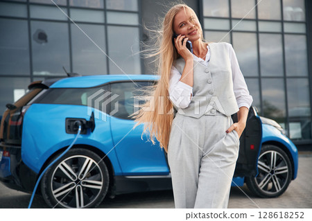 Using smartphone. Woman on the electric cars charge station at daytime Using smartphone. Woman on the electric cars charge station at daytime 128618252