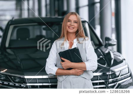 Black colored document in hands. Woman in formal clothes is in the car dealership Black colored document in hands. Woman in formal clothes is in the car dealership 128618266