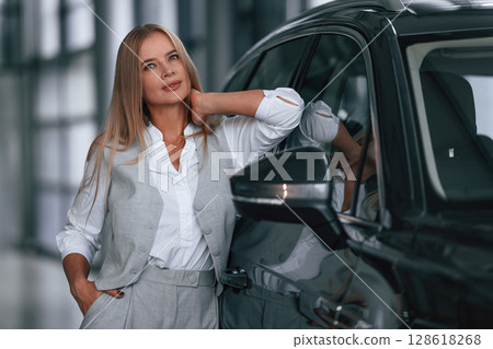 Leaning on the automobile. Woman in formal clothes is in the car dealership Leaning on the automobile. Woman in formal clothes is in the car dealership 128618268