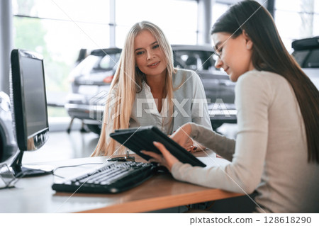 Holding tablet. Female manager is helping woman customer in the car dealership salon 128618290