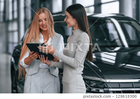 Standing and holding tablet. Female manager is helping woman customer in the car dealership salon 128618300
