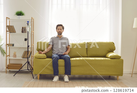 Young man receiving IV drip infusion and vitamin therapy sitting on the couch at home. Young man receiving IV drip infusion and vitamin therapy sitting on the couch at home. 128619714