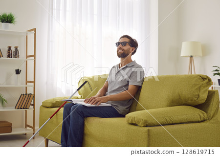 Blind Man Smiles While Reading Braille Book on Sofa 128619715