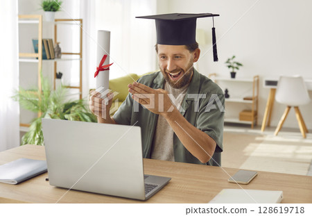 Young happy smiling graduate man having video call holding diploma in hands. Young happy smiling graduate man having video call holding diploma in hands. 128619718
